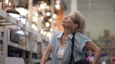 Yürüteç kullanan yaşlı bir kadın bir nalbur dükkanının koridorlarını araştırıyor. Yaşam alanını genişletmek için ev dekorasyonu malzemeleri arıyor..