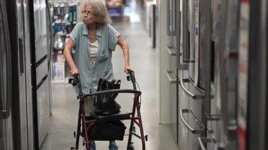 Yaşlı bir kadın, evini yenilemek için bir nalbur dükkanında malzeme ararken yürüteç kullanıyor, eşyalar ve malzemeler arasında geziniyor..