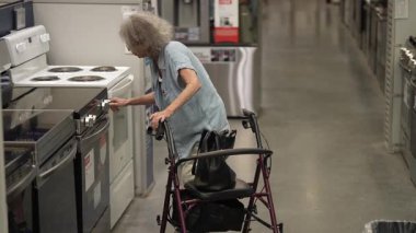 Yürüteç kullanan yaşlı bir kadın bir hırdavatçıda mutfak aletlerini dikkatle inceliyor, evini yenileme projesine odaklanıyor ve en iyi seçenekleri arıyor..
