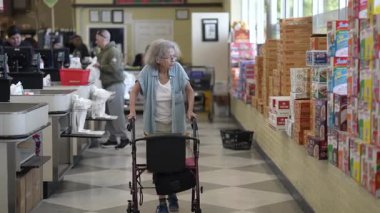 Winchester, VA, ABD - 07 22 2025: Engelli yaşlı bir kadın bakkaldan alışveriş yaparken raflardaki eşyaları kontrol ederek bir süpermarkette yürür. Arka planda diğer müşteriler görülebilir.