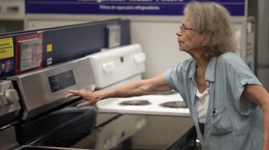 Yaşlı bir kadın, bir hırdavatçıda ev dekorasyonu ürünlerini incelerken yürüteç kullanıyor. İhtiyaçları için mutfak aletini dikkatlice inceliyor..