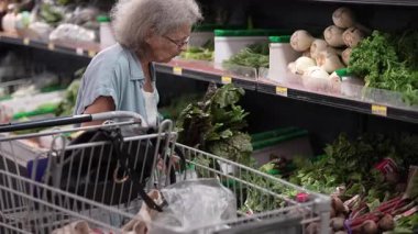 Yaşlı bir kadın markette taze sebzeleri karıştırıyor, yapraklı sebzeleri dikkatlice seçiyor ve gün boyunca alışveriş arabasına koyuyor..