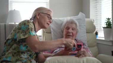 Gülümseyen hemşire yaşlı bir kadınla huzurevinde iletişim kurar, fotoğraf ve anılarını telefonla paylaşır, ziyaretleri sırasında iletişim ve hatıraları teşvik eder..