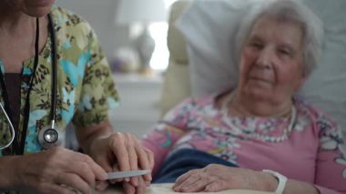 Hemşire, yaşlı bir kadının tırnaklarını nazikçe törpülerken tıbbi bir ortamda dinleniyor. Etkinlik, kıdemli hastalara gösterilen ilgi ve ilgiyi vurguluyor.