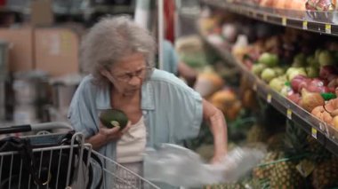 Yaşlı bir yaşlı kadın, çeşitli meyvelerle dolu bir marketten taze avokado seçiyor ve sağlıklı beslenmeye olan bağlılığını gösteriyor..