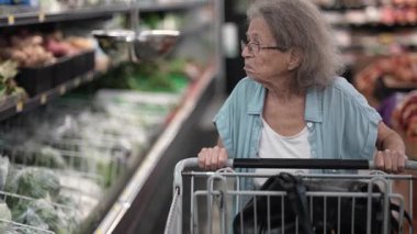 Yaşlı bir yaşlı kadın alışveriş arabasını öğleden sonra saatlerinde taze ürünlerle dolu bir mağazada market seçeneklerini düşünürken itiyor..