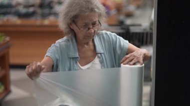 Yaşlı yaşlı bir yaşlı kadın iyi stoklanmış bir mağazada market alışverişi yaparken ürünleri dikkatle inceliyor. Eşyalarını seçmeye odaklanmış görünüyor..