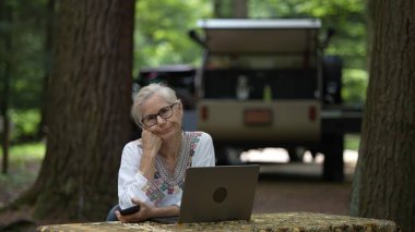 Huzurlu bir kamp alanında, olgun, emekli bir kadın dizüstü bilgisayarı ve akıllı telefonuyla meşgul oluyor, uzun ağaçlar ve rahat bir karavan ile çevrili..