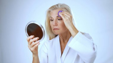 Olgun bir kadın, kaplıca elbisesi giyerken aynanın karşısına makyajını seve seve yapar..