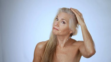 Olgun bir kadın spa tedavisinden zevk alırken neşeyle gülümser. Uzun saçları hafif ıslak ve yumuşak beyaz arka plan rahatlatıcı atmosfere katkı sağlıyor..