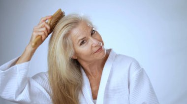 Beyaz spa bornozlu olgun bir kadın saçlarına nazikçe dokunurken sakin bir yüz ifadesi sergiliyor. Sakinleştirici bir ortamda mutluluk ve sükunet yayıyor..