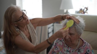 Olgun bir kadın, sıcak bir oturma odasında sıcak bir ziyaret sırasında sevgi ve ilgi göstererek yaşlı annesinin saçlarını nazikçe tarar..