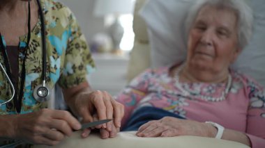 Hemşire, yaşlı bir kadının tırnaklarını nazikçe törpülerken tıbbi bir ortamda dinleniyor. Etkinlik, kıdemli hastalara gösterilen ilgi ve ilgiyi vurguluyor.