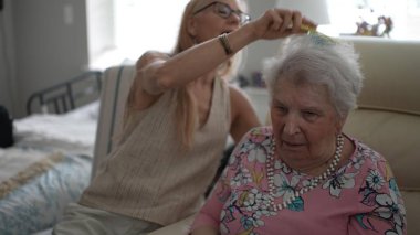 Olgun bir kadın, sıcak bir oturma odasında yaşlı annesinin saçlarını nazikçe tarar, samimi bir ziyaret sırasında bağlarının altını çizer..