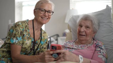 Hemşire ve yaşlı bir kadının portresi sıcak bir şekilde etkileşime geçiyor, yaşlılar yurduna yapılan bir ziyarette telefon kullanarak anılarını ve bilgilerini paylaşıyor..