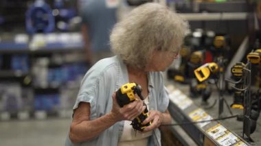 Yaşlı bir kadın elinde bir yürüteçle bir hırdavatçıda geziniyor. Ev yenileme projesi için gerekli alet ve gereçleri dikkatlice inceliyor. Odaklanmış ve kararlı görünüyor..