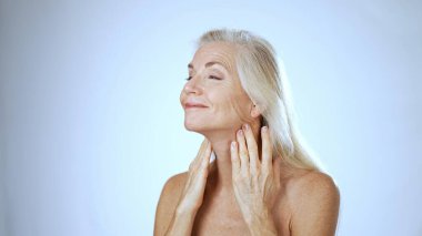 Uzun, açık renk saçlı olgun bir kadın, sakin, beyaz bir arka planda spa tedavisinden zevk alırken mutlu bir şekilde gülümsüyor. Mutluluğu huzur ve rahatlık veriyor..