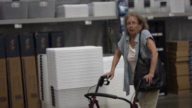 Yaşlı bir kadın bir hırdavatçıda boya seçeneklerini araştırıyor, destek için bir yürüteç kullanıyor. Ev yenileme projesi için renkleri inceliyor..