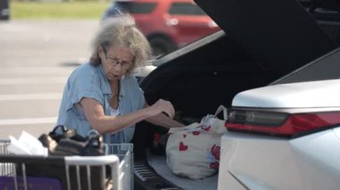 Yaşlı bir kadın marketten alışveriş yaparken görülüyor. Eşyalarını dikkatli bir şekilde açık gökyüzünün altındaki otoparktaki arabasına taşıyor..