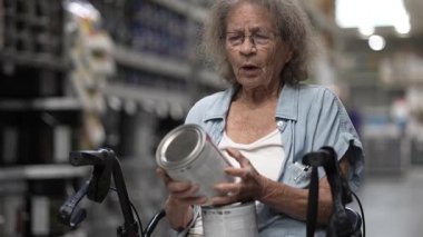 Gri saçlı yaşlı bir kadın yürüteçle otururken boya kutularını inceliyor ve bir nalbur dükkanındaki ev yenileme projesi için renk seçmeye odaklanıyor..