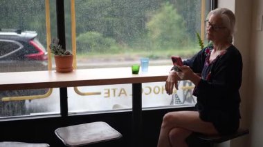 Gülümseyen olgun bir kadının portresi kafeteryada oturuyor, kahve içiyor ve bilgisayarına ve telefonuna odaklanıyor. Atmosfer yağmurlu bir manzarayla sıcak..