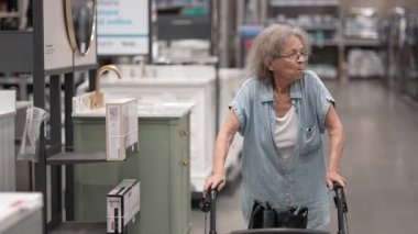 Yürüteci olan yaşlı bir kadın, restorasyon projesi için malzeme seçerek, geniş bir hırdavatçıda ev dekorasyonu malzemeleri arıyor..