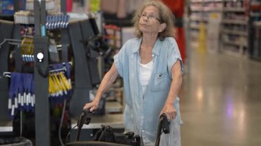 Yaşlı, yaşlı bir kadın, bir nalbur dükkanının kasasında yürüteç kullanırken kendini kontrol ediyor. Alışverişine odaklanmış görünüyor..