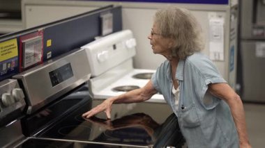 Yürüteç kullanan yaşlı bir kadın hırdavatçıdaki eşyaları elden geçiriyor. Ev projesi için seçenekleri düşünerek yeni bir sobayı inceliyor..