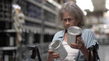 Yaşlı bir yaşlı kadın, Walker 'la hırdavatçıda alışveriş yaparken iki boya kutusu arasında seçim yapıyor. Ev projesi için seçenekleri dikkatlice inceliyor..