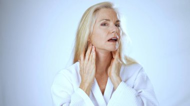 Beyaz cüppeli olgun bir kadın spa 'da mutlu bir şekilde gülümseyerek rahatlatıcı bir anın tadını çıkarıyor. Huzurlu ortam onun rahatlama ve mutluluk duygusunu arttırır..