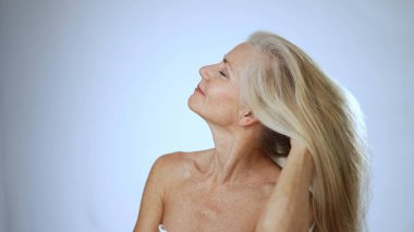 Kaplıcadaki olgun bir kadın uzun, dalgalı saçlarını yumuşak beyaz arka planda şekillendirirken neşeli bir şekilde gülümsüyor. Mutluluk ve sükunet saçar..