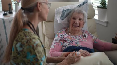 Bir hemşire, rutin bir ziyaret sırasında hayati organlarını kontrol ederken gülümseyen mutlu ve yaşlı bir kadınla hoş bir sohbet eder..