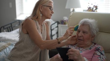 Olgun bir kadın yaşlı annesinin saçlarını dikkatlice tarar ve ziyaretleri sırasında yumuşak doğal ışıkla dolu sıcak bir odada huzurlu bir an yaratır..