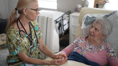 Bir hemşire yaşlı bir kadının elini tutarak huzur verici bir ortam yaratmak için huzur verici bir huzurevinde dostça bir konuşma yapar..