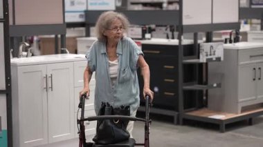 Yaşlı bir kadın, bir hırdavatçıda çeşitli ev yenileme malzemelerini karıştırırken yürüteç kullanıyor. Alışveriş tecrübesiyle meşgul görünüyor..
