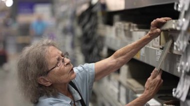 Yürüteç kullanan yaşlı bir kadın, hırdavatçıda alışveriş yaparken ev dekorasyonu için malzemeleri dikkatlice seçiyor. Seçeneklerle dolu yoğun bir gün..