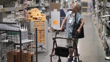 Kıdemli bir kadın hırdavatçıda ev tadilatı için yürüteç kullanıyor. Koridordaki seçeneklerini düşünürken odaklanmış görünüyor..
