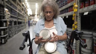 Yürüyen tekerlekli sandalyedeki yaşlı bir kadın bir hırdavatçıda boya kutularını inceliyor, gelecek dekorasyon projesi için doğru renkleri seçmeye odaklanmış..