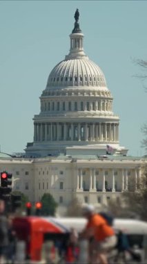 ABD Kongre Binası 'nın dikey videosu açık mavi gökyüzünün altında duruyor. Arabalar caddelerde geziniyor ve bayraklar bölgeyi süslüyor, hareketli bir atmosfer yaratıyor..
