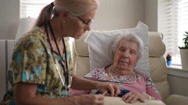 Hemşire, yaşlı bir kadının tırnaklarını rahatça törpülüyor. İlgili etkileşim, ziyaret sırasında bir anlık şefkat ve kişisel ilgiyi vurgular..