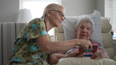Gülümseyen hemşire yaşlı bir kadınla huzurevinde iletişim kurar, fotoğraf ve anılarını telefonla paylaşır, ziyaretleri sırasında iletişim ve hatıraları teşvik eder..
