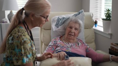 Bir hemşire yaşlı bir kadının oksijen seviyesini oksitölçer kullanarak dikkatle izler. Parlak bir odadalar, yaşlılar için sağlık hizmetleri ve desteklerini ön plana çıkarıyorlar..