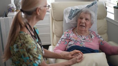 Huzurevindeki yaşlı kadın el ele tutuşurken bir hemşireyle dostça sohbet ediyor. Atmosfer sıcak ve destekleyici..