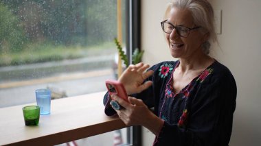 Olgun bir kadın kafeterya masasında oturup kahvesini yudumluyor ve telefonuyla görüntülü bir sohbete girişiyor. Yağmurlu bir arka planda işine odaklanıyor..