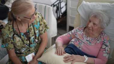 Neşeli bir hemşire yaşlı bir kadınla konuşurken huzur ve refahını sağlamak için huzurevinde yaşam belirtilerini kontrol eder..