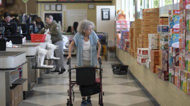 Engelli yaşlı bir kadın yürüteci ile süpermarkette geziniyor, alışveriş yaparken raflardaki eşyaları kontrol ediyor. Arka planda diğer müşteriler görülebilir.