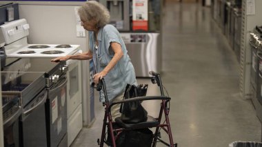 Yürüteç kullanan yaşlı bir kadın bir hırdavatçıda mutfak aletlerini dikkatle inceliyor, evini yenileme projesine odaklanıyor ve en iyi seçenekleri arıyor..