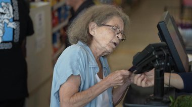 Yaşlı bir kadın kasada, bakkaliyesinin parasını ödüyor. O, yoğun alışveriş gününde kasiyerle etkileşime girerken odaklanmış görünüyor..