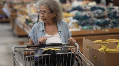 Gri saçlı yaşlı bir kadın alışveriş arabasını iterken kalabalık bir markette taze yiyecek seçiyor. Listesine ve yakındaki ürünlere odaklanıyor..
