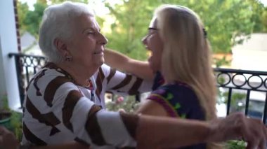 Gülerken, yaşlı bir kadın ve genç bir kadın balkonda birbirlerine sarılıyorlar..